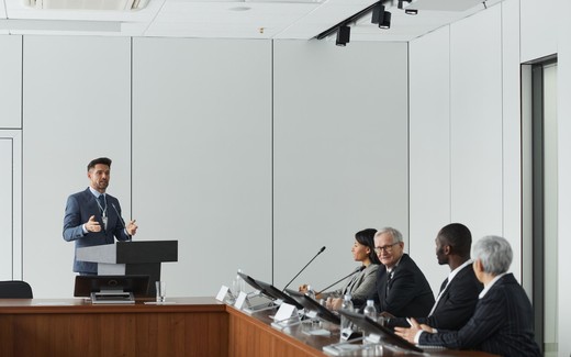 A speaker addresses a seated panel of four across a conference table