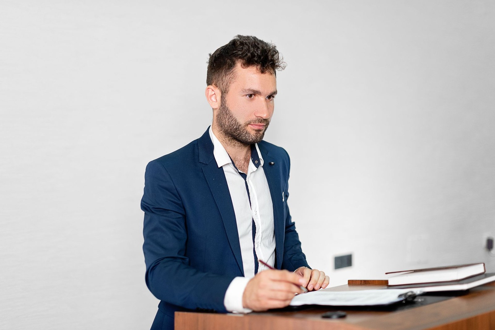 A presenter in a dark suit reviews prepared notes at a lectern, pen in hand, before a formal session
