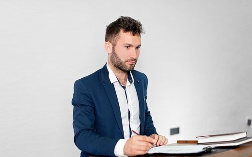A presenter in a dark suit reviews prepared notes at a lectern, pen in hand, before a formal session