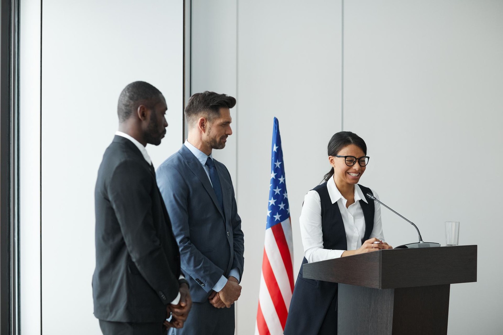 A woman in glasses smiles while speaking from a lectern, with an American flag and two colleagues standing nearby