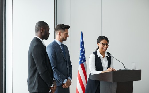 A woman in glasses smiles while speaking from a lectern, with an American flag and two colleagues standing nearby
