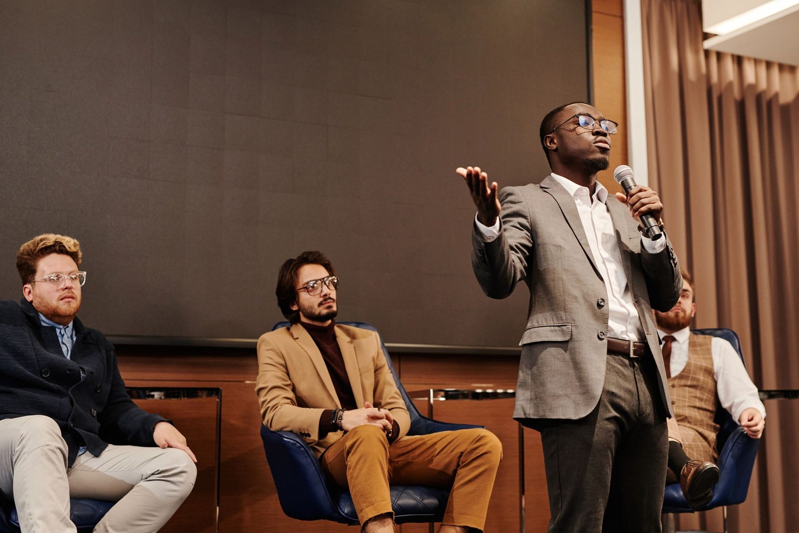 A panelist gestures while speaking into a microphone as fellow panelists listen at a discussion-format meeting