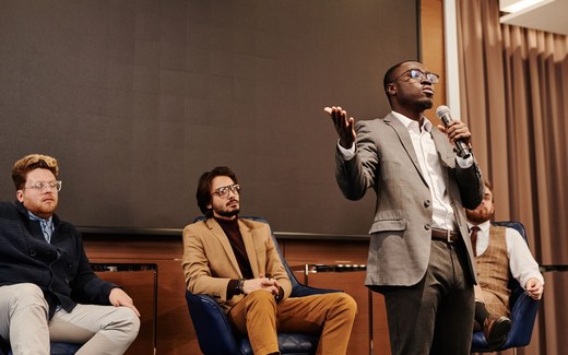 A panelist gestures while speaking into a microphone as fellow panelists listen at a discussion-format meeting