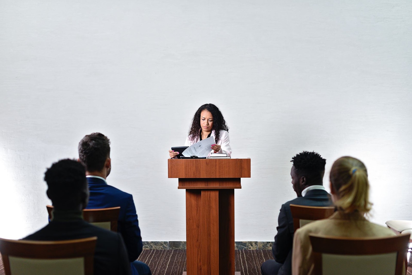 A speaker at a lectern reads from prepared remarks while an audience listens from rows of chairs