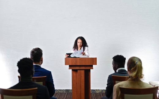 A speaker at a lectern reads from prepared remarks to a seated audience