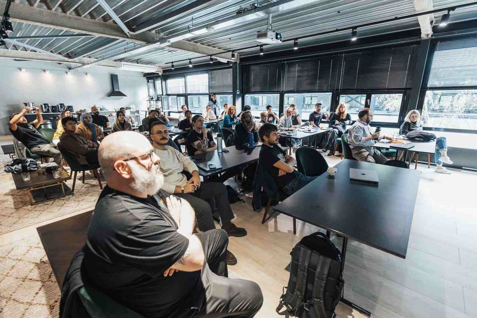 Attendees seated at tables in a bright, modern meeting space listening to a presentation