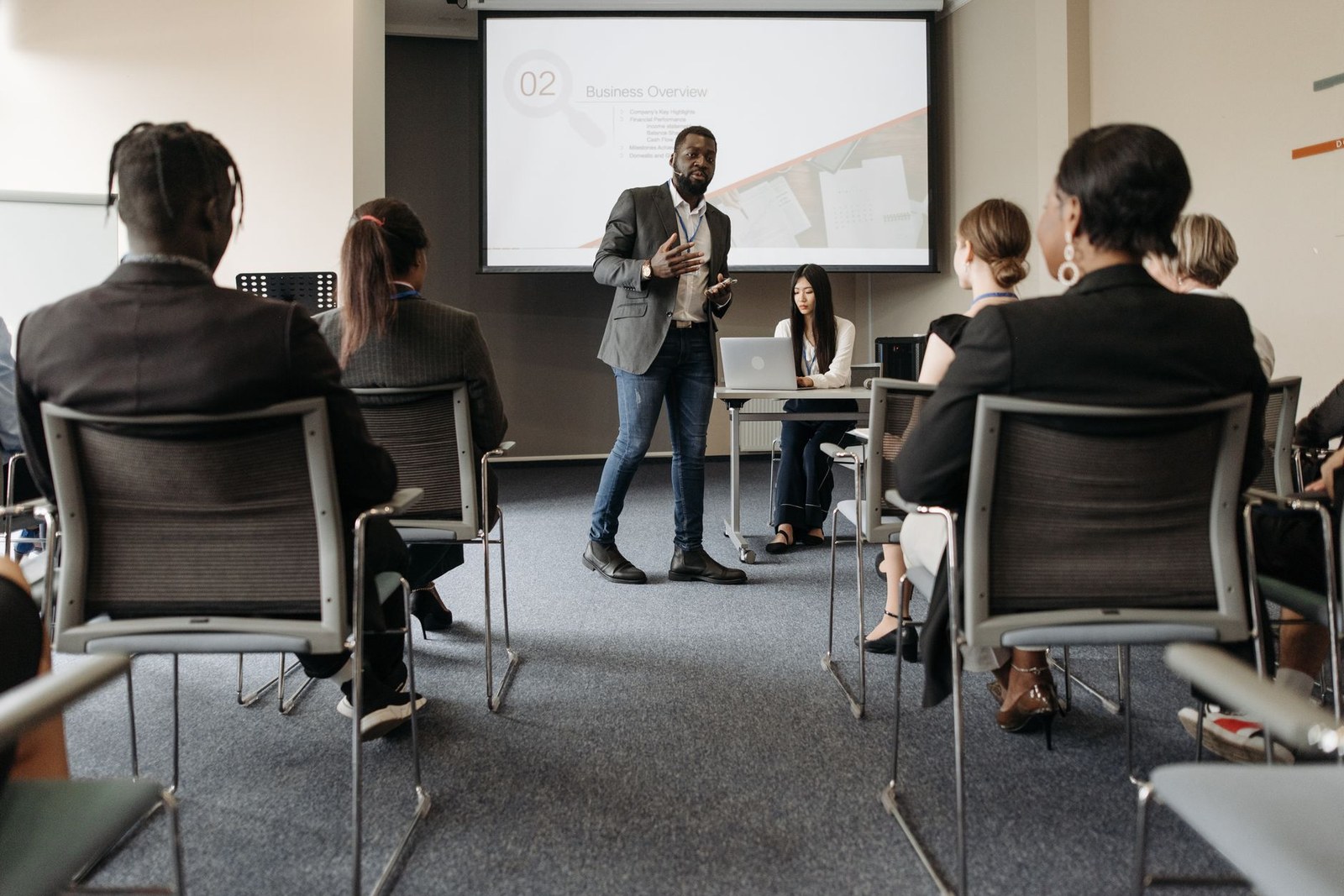 A presenter speaks in front of a screen to a seated audience in a training room
