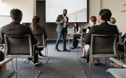 A presenter speaks in front of a screen to a seated audience in a training room