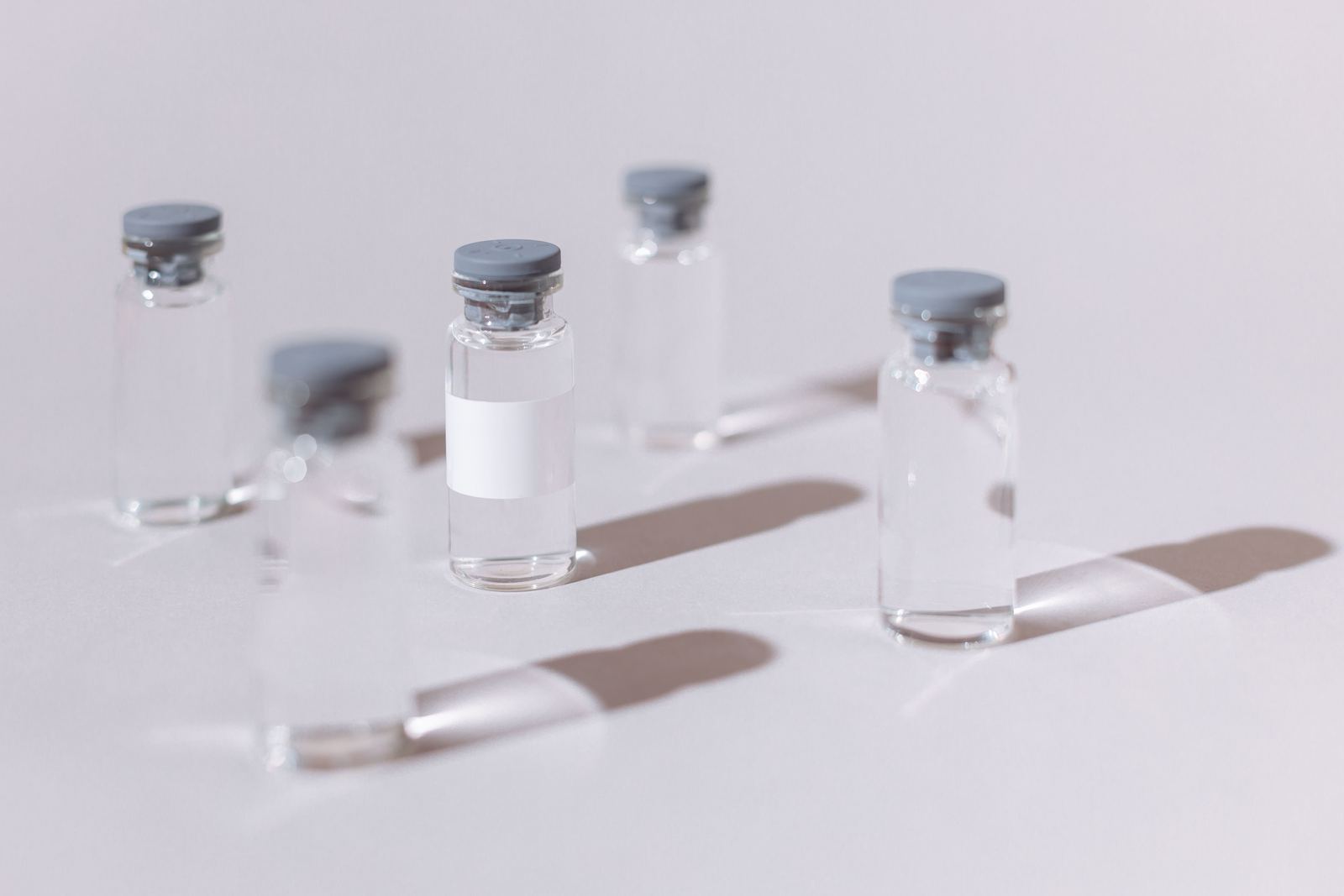Clear glass injection vials with grey stoppers casting long shadows on a white surface