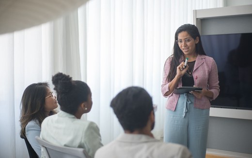 A presenter in a pink blazer addresses colleagues seated around her during a workshop session