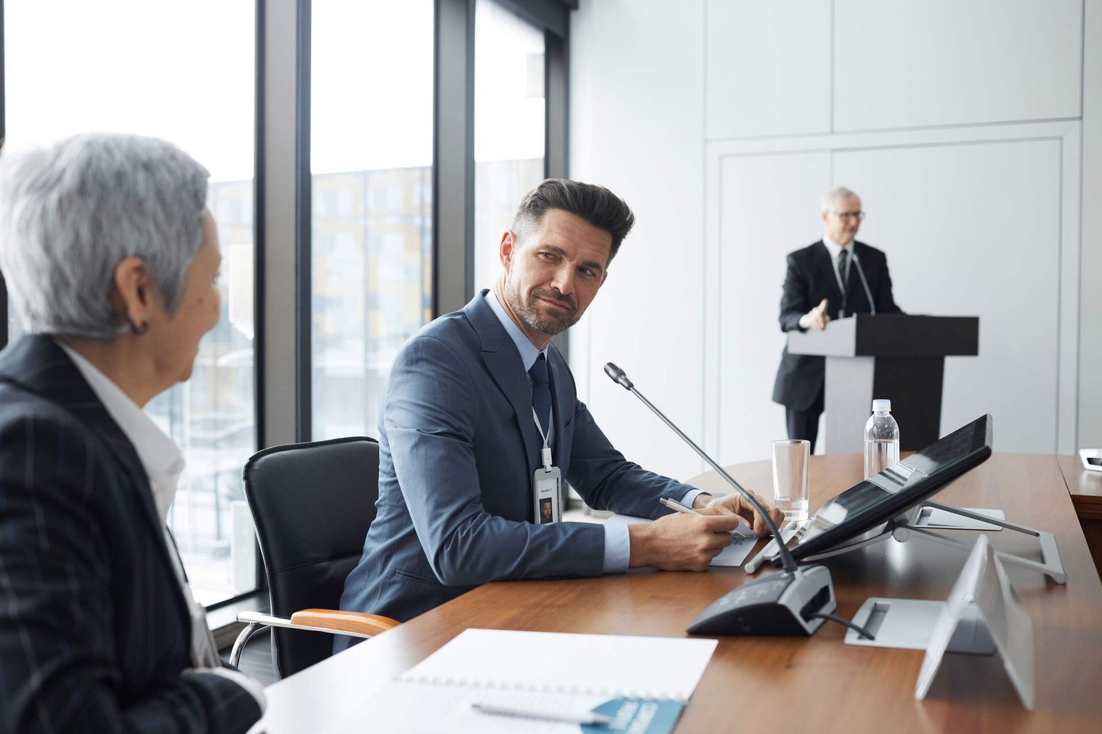 A delegate seated at a conference table glances toward the camera while a speaker addresses attendees from a podium in the background