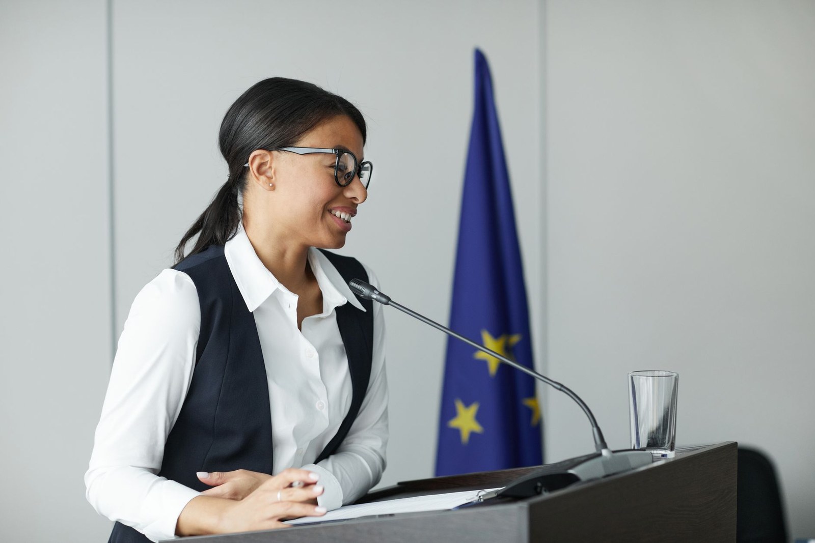 A regulatory professional smiling while speaking from a lectern, with the European Union flag behind her