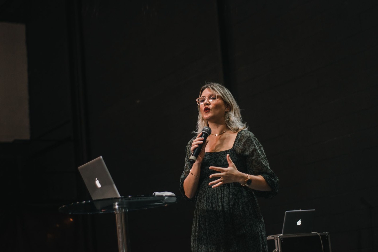 A woman in glasses speaking into a handheld microphone while presenting from a stage