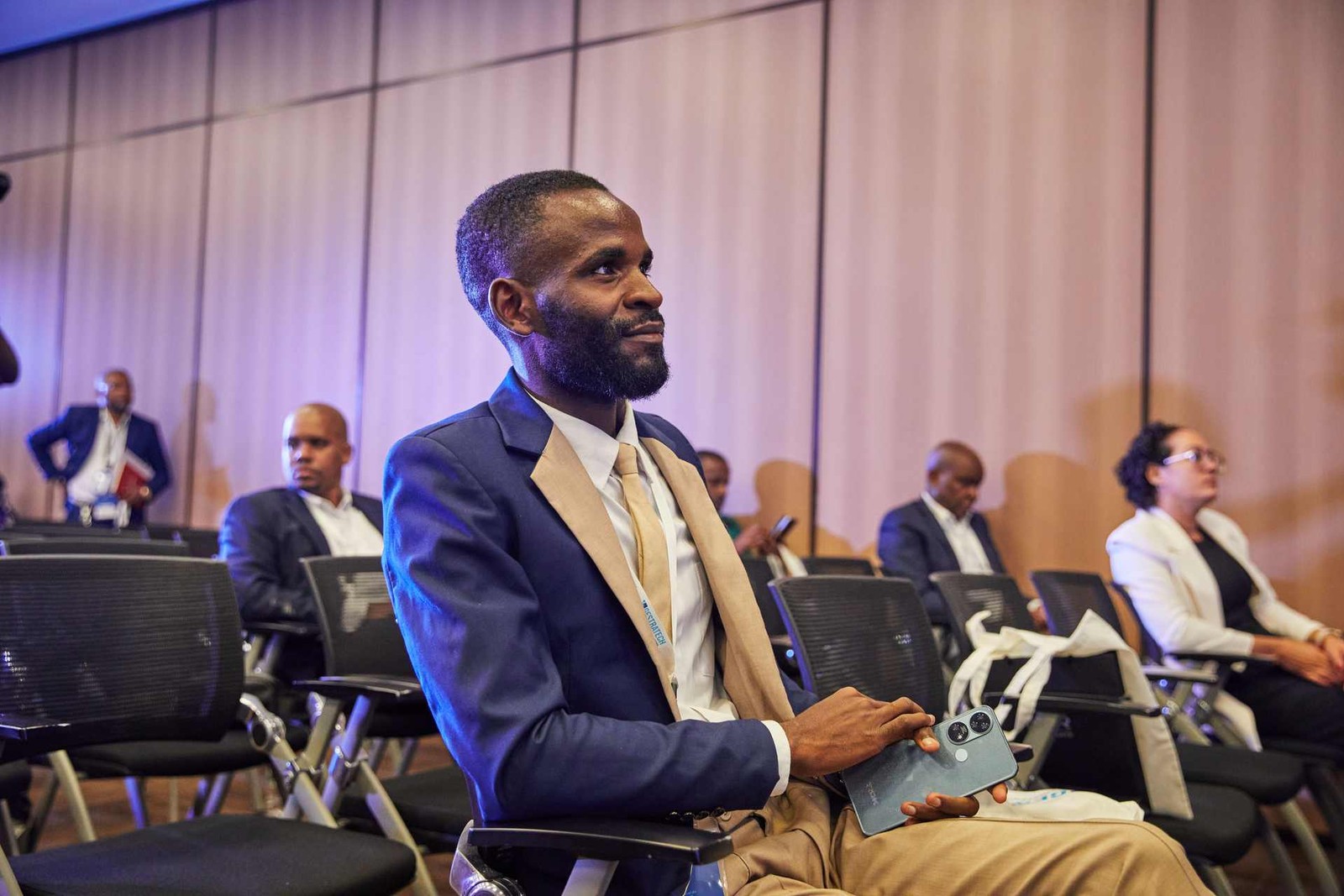 A man in a suit and tan tie sits attentively among rows of conference attendees, holding a phone