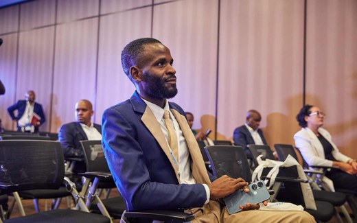 A man in a suit and tan tie sits attentively among rows of conference attendees, holding a phone