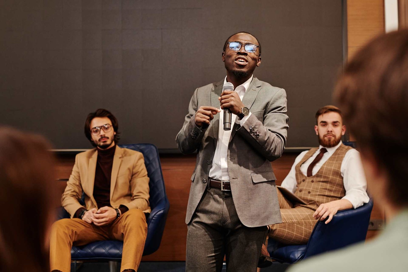 A speaker addressing a panel discussion, flanked by two seated panelists, in a bright modern conference room