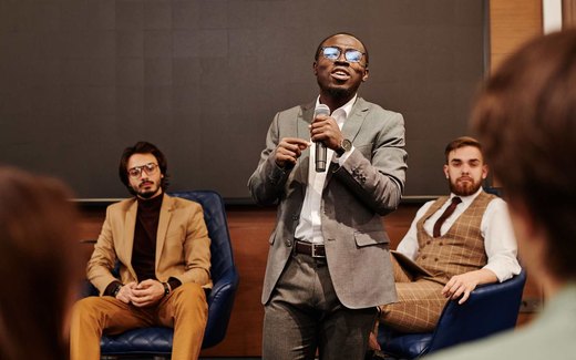 A panelist speaking into a microphone while two fellow panelists listen, in a bright conference room