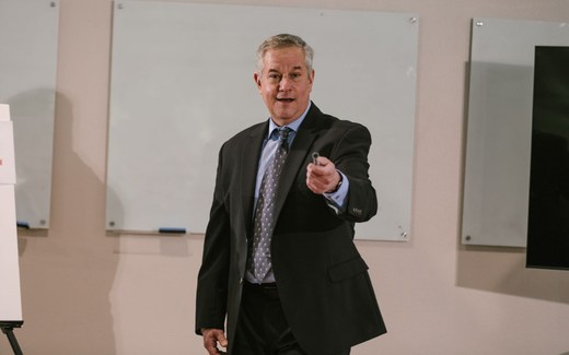 A presenter in a suit gestures while speaking to a small group in a training room